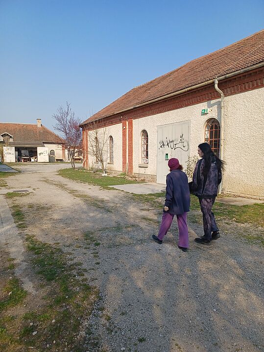 Zwei Personen gehen auf einem Kiesweg neben einem Gebäude mit roten Ziegeln und Graffiti an der Tür