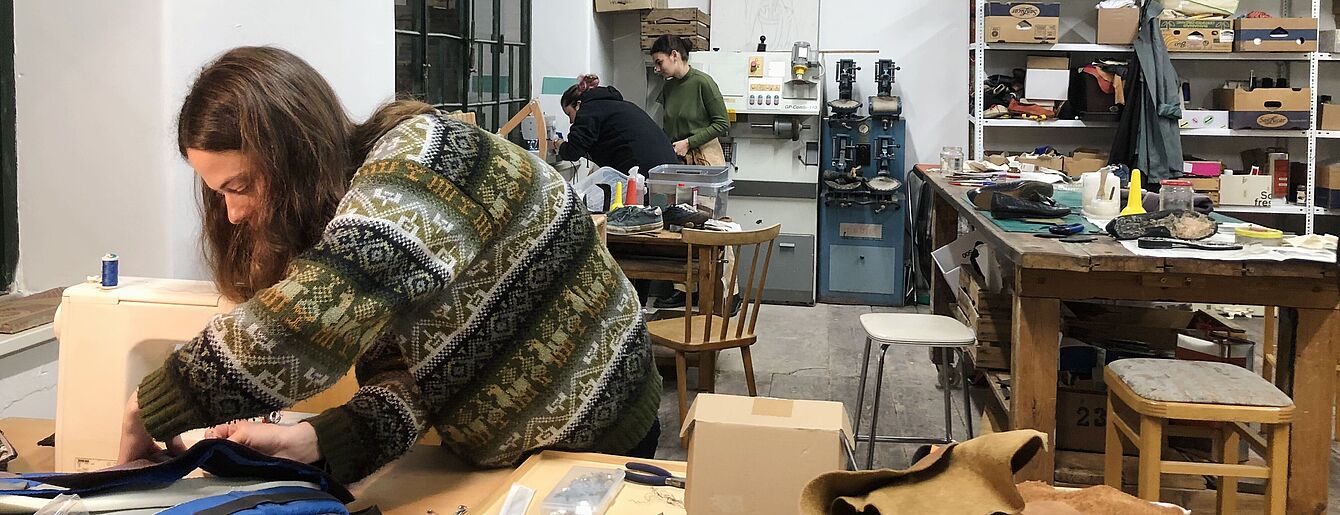 Person arbeitet an Nähmaschine in Werkstatt mit Tischen, Werkzeugen und Regalen im Reparaturcafe