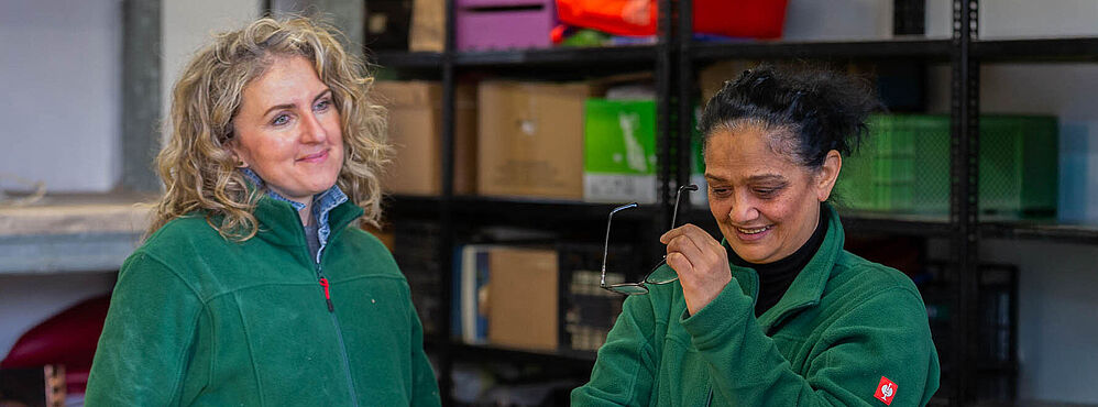 Foto: Susanne Senekowitsch Zwei Personen in grünen Jacken stehen vor Regalen mit bunten Aufbewahrungsboxen in einem Lagerraum.