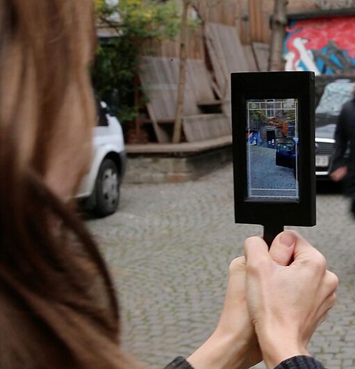 Eine Person hält ein Smartphone mit sichtbarem Kamerabild auf eine Gruppe von vier Personen, die auf einem gepflasterten Platz laufen