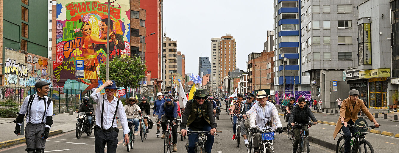 Fahrradfahrer*innen auf einer breiten Straße, im Hintergrund sieht man ein riesiges farbenfrohes Mural auf einer Feuermauer