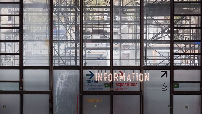 Große Glasfenster mit sichtbarem Gerüst dahinter, darauf leuchtende Schrift 'INFORMATION' und Richtungspfeile zu Museum, Ausstellungen und Kino.