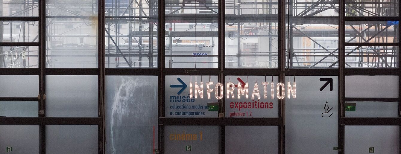 Große Glasfenster mit sichtbarem Gerüst dahinter, darauf leuchtende Schrift 'INFORMATION' und Richtungspfeile zu Museum, Ausstellungen und Kino.