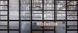 Große Glasfenster mit sichtbarem Gerüst dahinter, darauf leuchtende Schrift 'INFORMATION' und Richtungspfeile zu Museum, Ausstellungen und Kino.