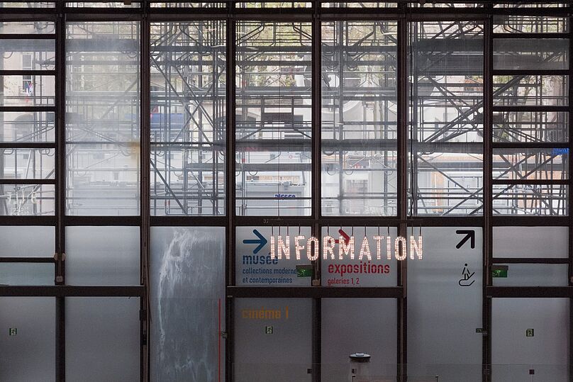 Große Glasfenster mit sichtbarem Gerüst dahinter, darauf leuchtende Schrift 'INFORMATION' und Richtungspfeile zu Museum, Ausstellungen und Kino.