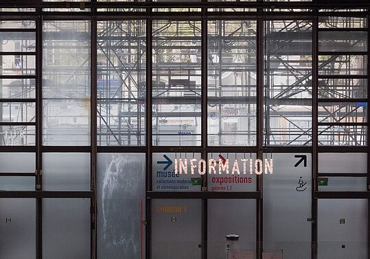Große Glasfenster mit sichtbarem Gerüst dahinter, darauf leuchtende Schrift 'INFORMATION' und Richtungspfeile zu Museum, Ausstellungen und Kino.