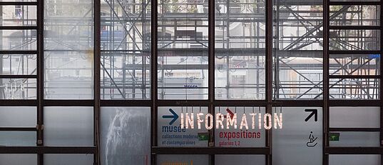 Große Glasfenster mit sichtbarem Gerüst dahinter, darauf leuchtende Schrift 'INFORMATION' und Richtungspfeile zu Museum, Ausstellungen und Kino.