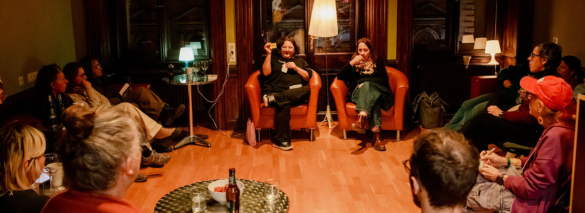 Mehrere Personen sitzen in einem Raum mit Holzfußboden, zwei Personen auf Sesseln in der Mitte, andere auf Stühlen im Halbkreis, Tisch mit Getränken im Vordergrund.