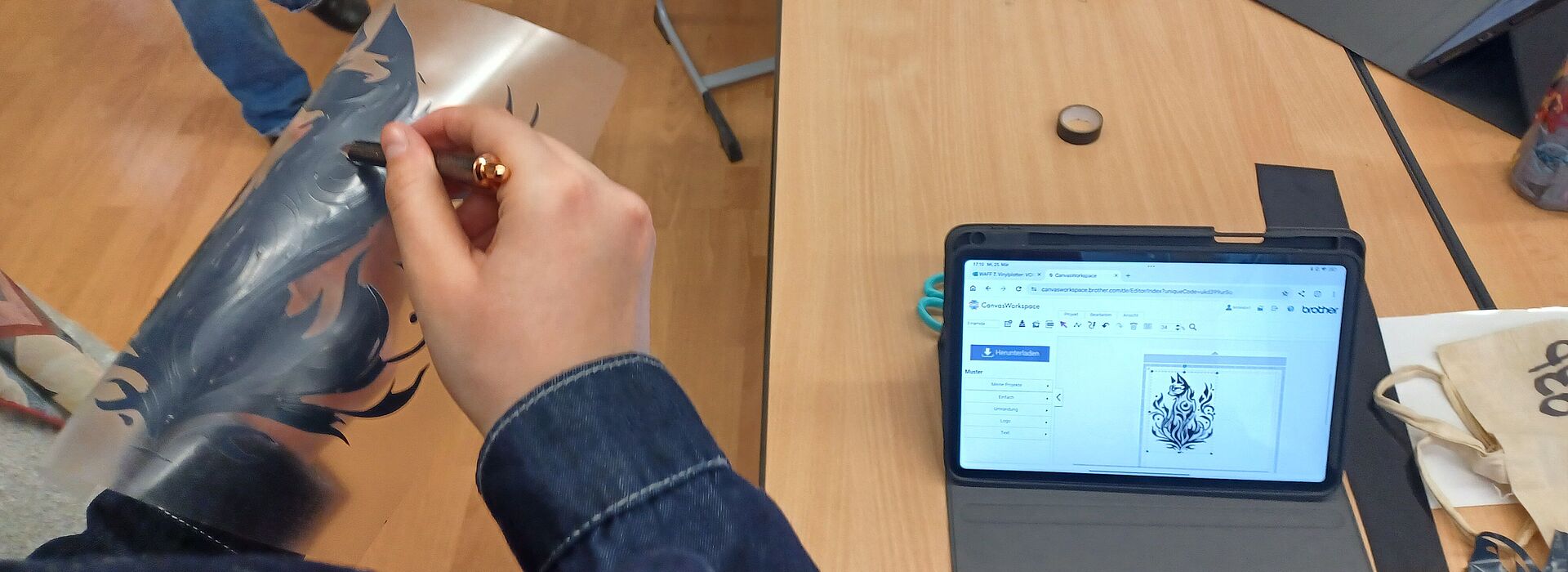 Zwei Personen halten eine Schablone mit einem Flammenmotiv über einem Tisch mit Tablet, Laptop und Wasserflasche.