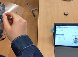 Zwei Personen halten eine Schablone mit einem Flammenmotiv über einem Tisch mit Tablet, Laptop und Wasserflasche.