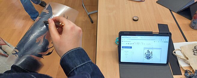 Zwei Personen halten eine Schablone mit einem Flammenmotiv über einem Tisch mit Tablet, Laptop und Wasserflasche.
