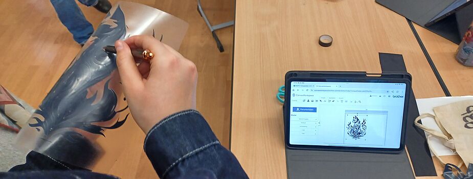 Zwei Personen halten eine Schablone mit einem Flammenmotiv über einem Tisch mit Tablet, Laptop und Wasserflasche.