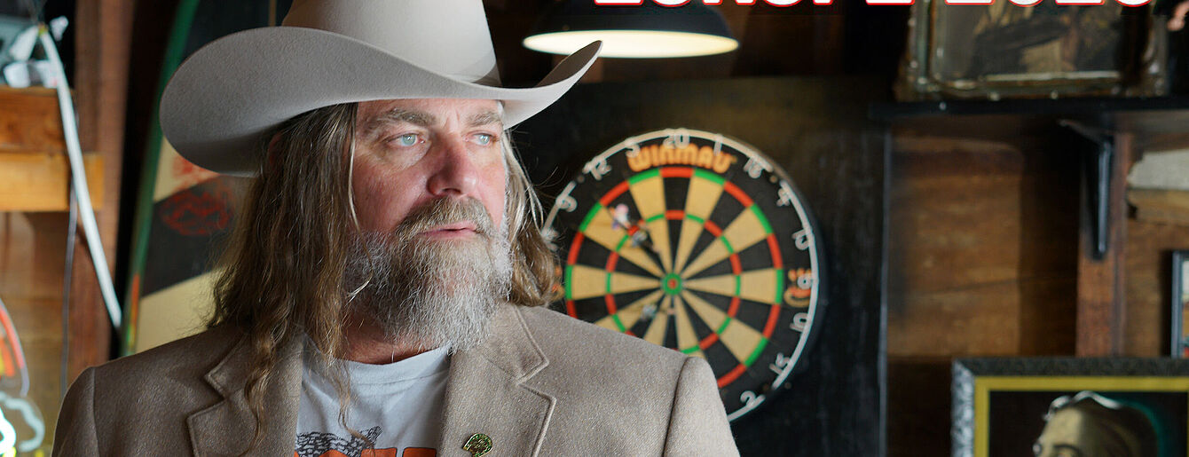 Person mit langem Haar und Bart trägt Cowboyhut und braunen Blazer vor Dartscheibe in rustikalem Raum