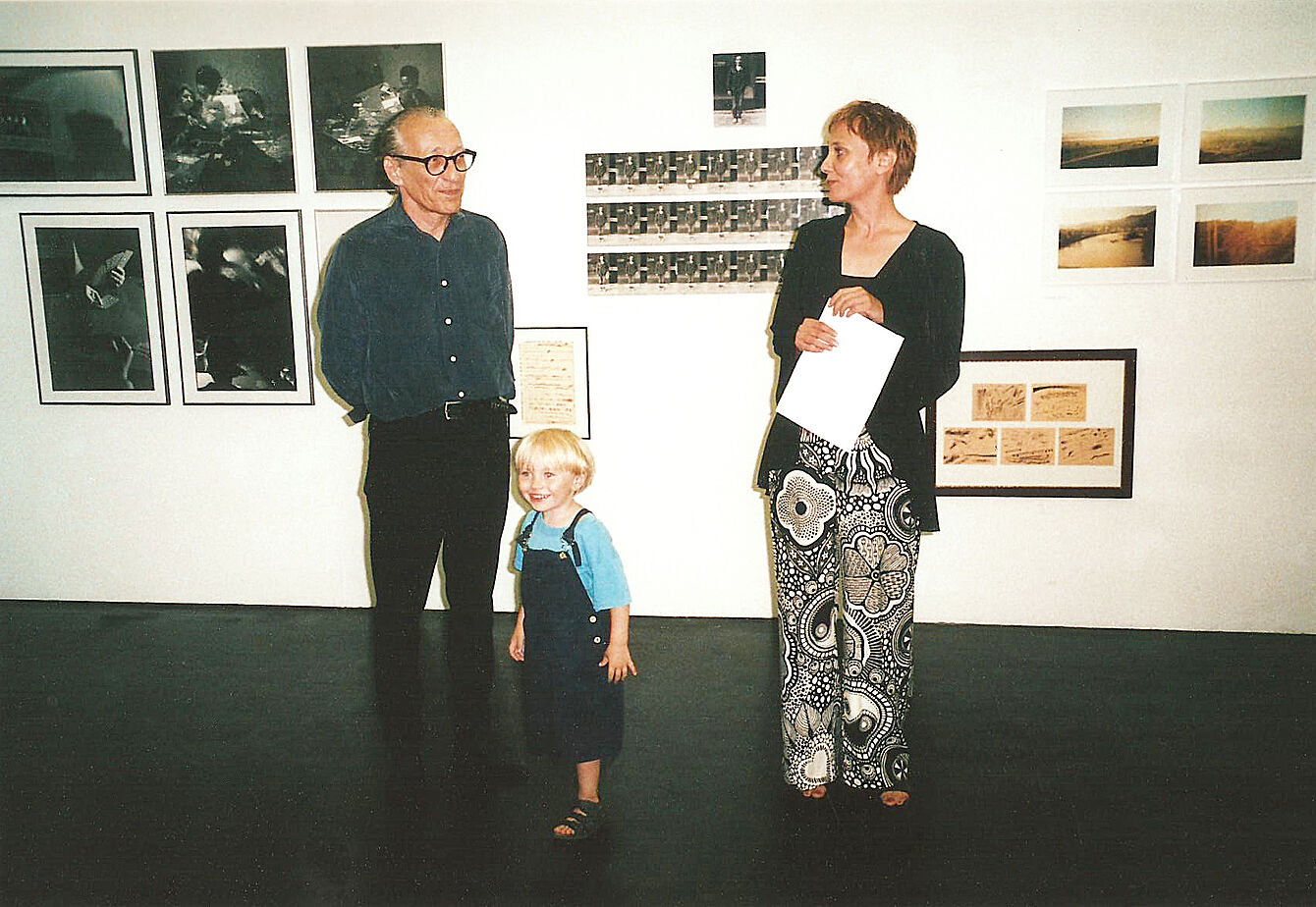 Drei Personen stehen in einem Ausstellungsraum mit schwarz-weißen Fotografien und Bildern an der Wand.