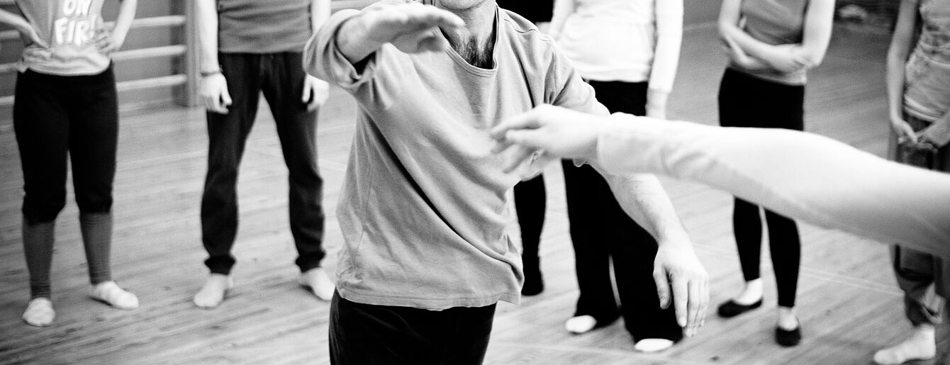 Person in Bewegung vor einer Gruppe von Menschen in einem Tanz- oder Theaterraum mit Holzboden