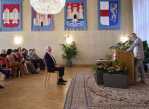 Mann am Rednerpult in festlichem Saal mit Publikum auf Stühlen und Wappen an der Wand