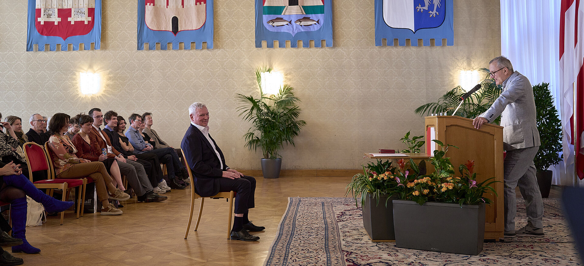 Mann am Rednerpult in festlichem Saal mit Publikum auf Stühlen und Wappen an der Wand