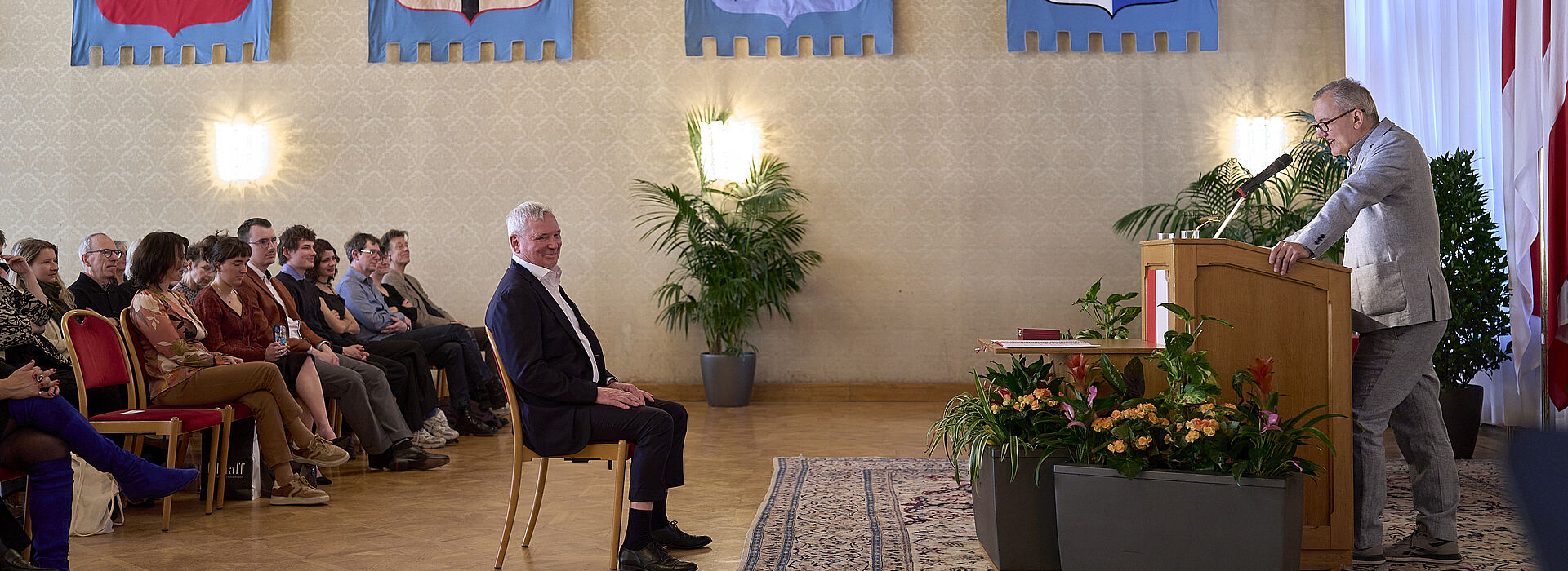 Mann am Rednerpult in festlichem Saal mit Publikum auf Stühlen und Wappen an der Wand