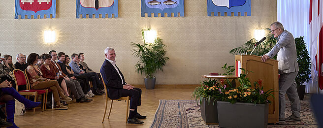 Mann am Rednerpult in festlichem Saal mit Publikum auf Stühlen und Wappen an der Wand