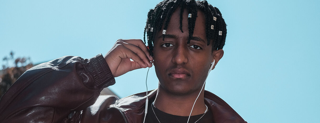 Porträt einer jungen Person mit kurzen Dreadlocks, die Kopfhörer trägt und eine Hand zum Ohr hebt