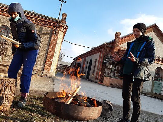 Zwei Personen in Winterkleidung stehen an einem Feuer in einer runden Feuerschale vor Backsteingebäuden