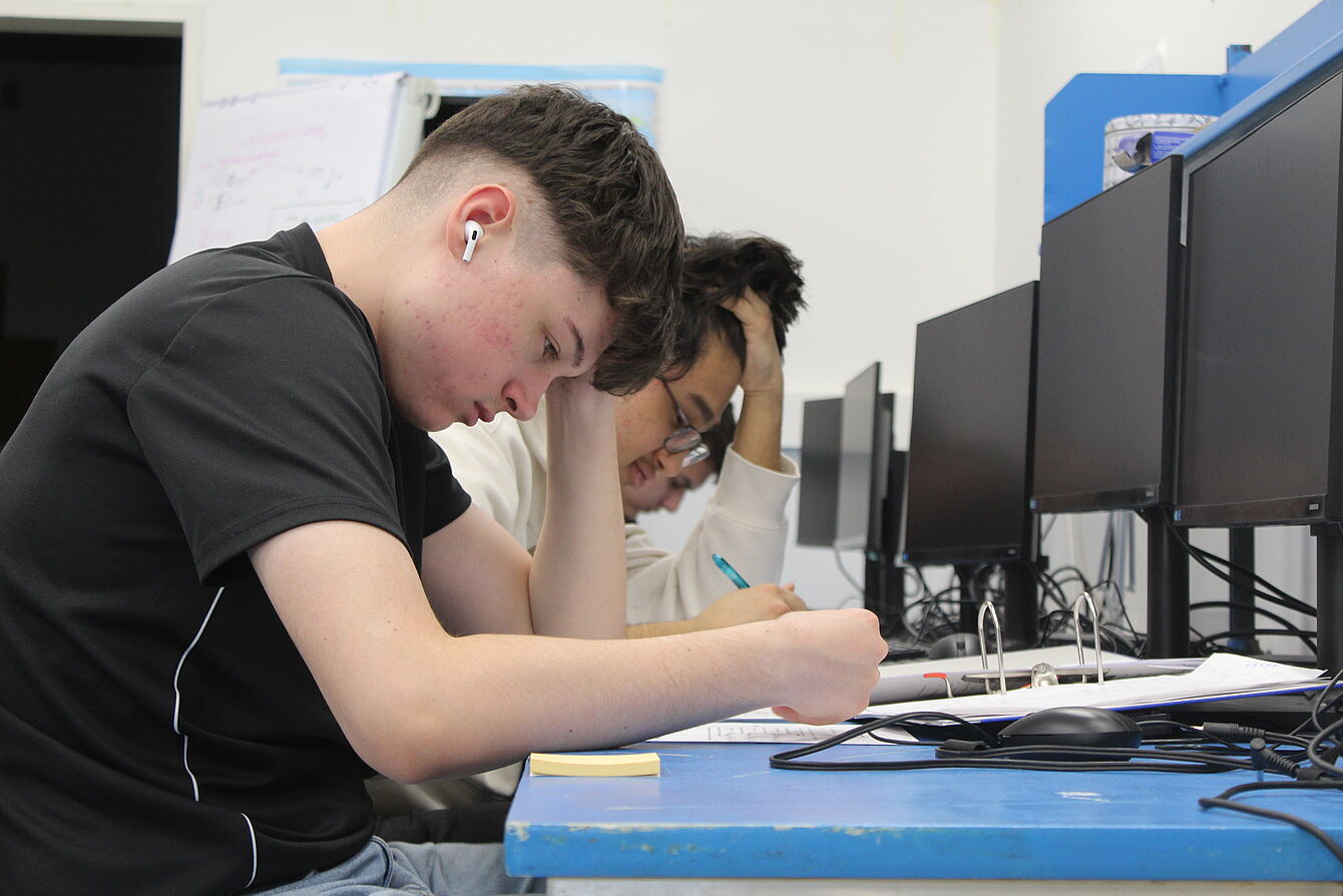 Zwei Jugendliche sitzen vor Computern und lesen sich ausgedruckte Unterlagen durch 