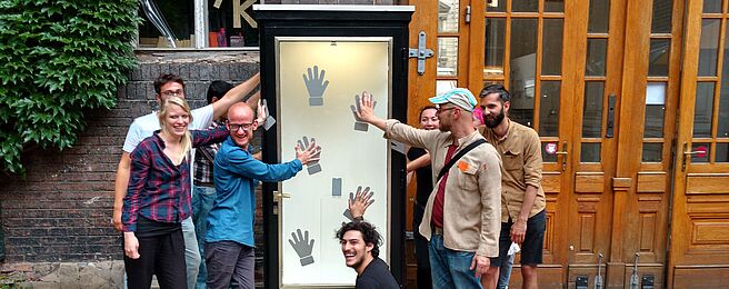 Gruppe von acht Personen vor einem hohen, schmalen Schaukasten mit mehreren schwarzen Handabdrücken auf weißem Hintergrund, einige Personen berühren die Handabdrücke.