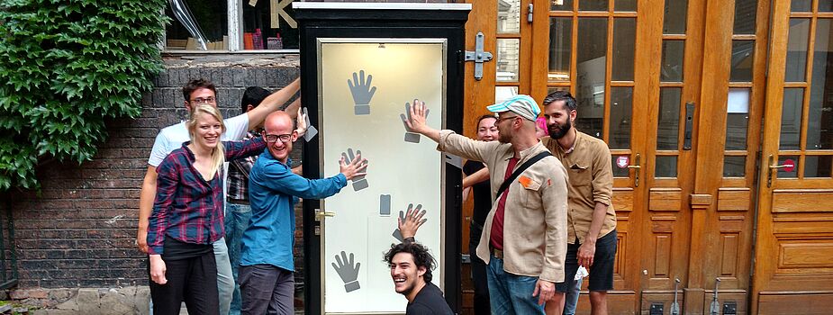 Gruppe von acht Personen vor einem hohen, schmalen Schaukasten mit mehreren schwarzen Handabdrücken auf weißem Hintergrund, einige Personen berühren die Handabdrücke.