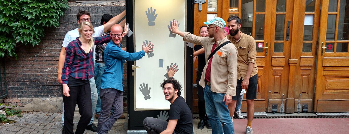 Menschen stehen um die Kunstzelle. Jede Person hat eine Hand an ihren Wänden.