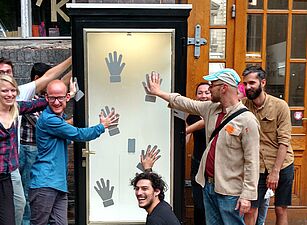 Gruppe von acht Personen vor einem hohen, schmalen Schaukasten mit mehreren schwarzen Handabdrücken auf weißem Hintergrund, einige Personen berühren die Handabdrücke.