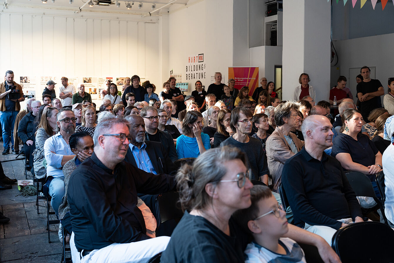 Publikum unterschiedlichen Alters sitzt in einem hellen Raum und blickt nach vorne