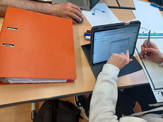 Zwei Personen an einem Tisch, eine zeigt auf ein Tablet mit einer Zeichnung, daneben ein großer oranger Ordner und Schreibutensilien