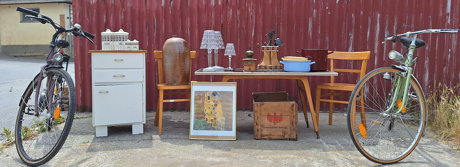 Zwei Fahrräder flankieren einen Tisch mit Stühlen, einer Kommode, Lampen, einem Bild und einer Holzkiste vor einer roten Wellblechwand.