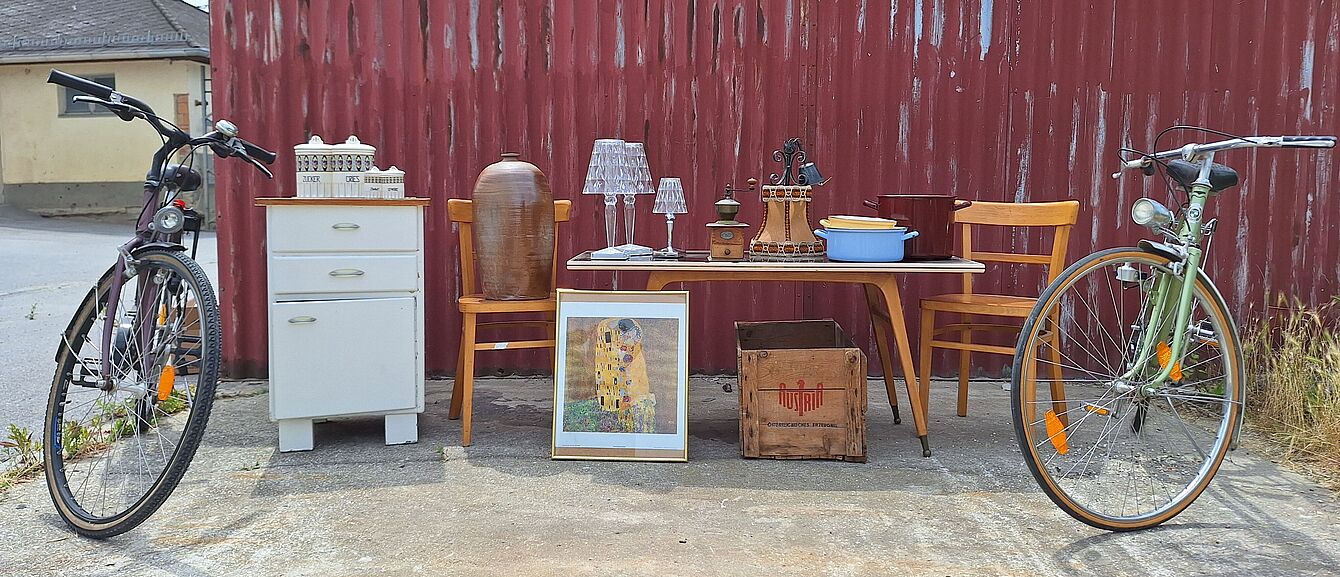 Zwei Fahrräder flankieren einen Tisch mit Stühlen, einer Kommode, Lampen, einem Bild und einer Holzkiste vor einer roten Wellblechwand.