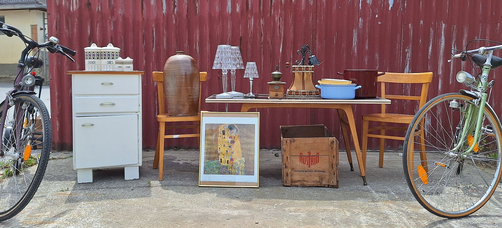 Zwei Fahrräder flankieren einen Tisch mit Stühlen, einer Kommode, Lampen, einem Bild und einer Holzkiste vor einer roten Wellblechwand.