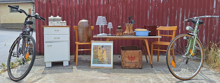 Zwei Fahrräder flankieren einen Tisch mit Stühlen, einer Kommode, Lampen, einem Bild und einer Holzkiste vor einer roten Wellblechwand.