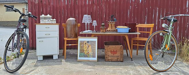 Foto: Lilly Kolb Zwei Fahrräder flankieren einen Tisch mit Stühlen, einer Kommode, Lampen, einem Bild und einer Holzkiste vor einer roten Wellblechwand.