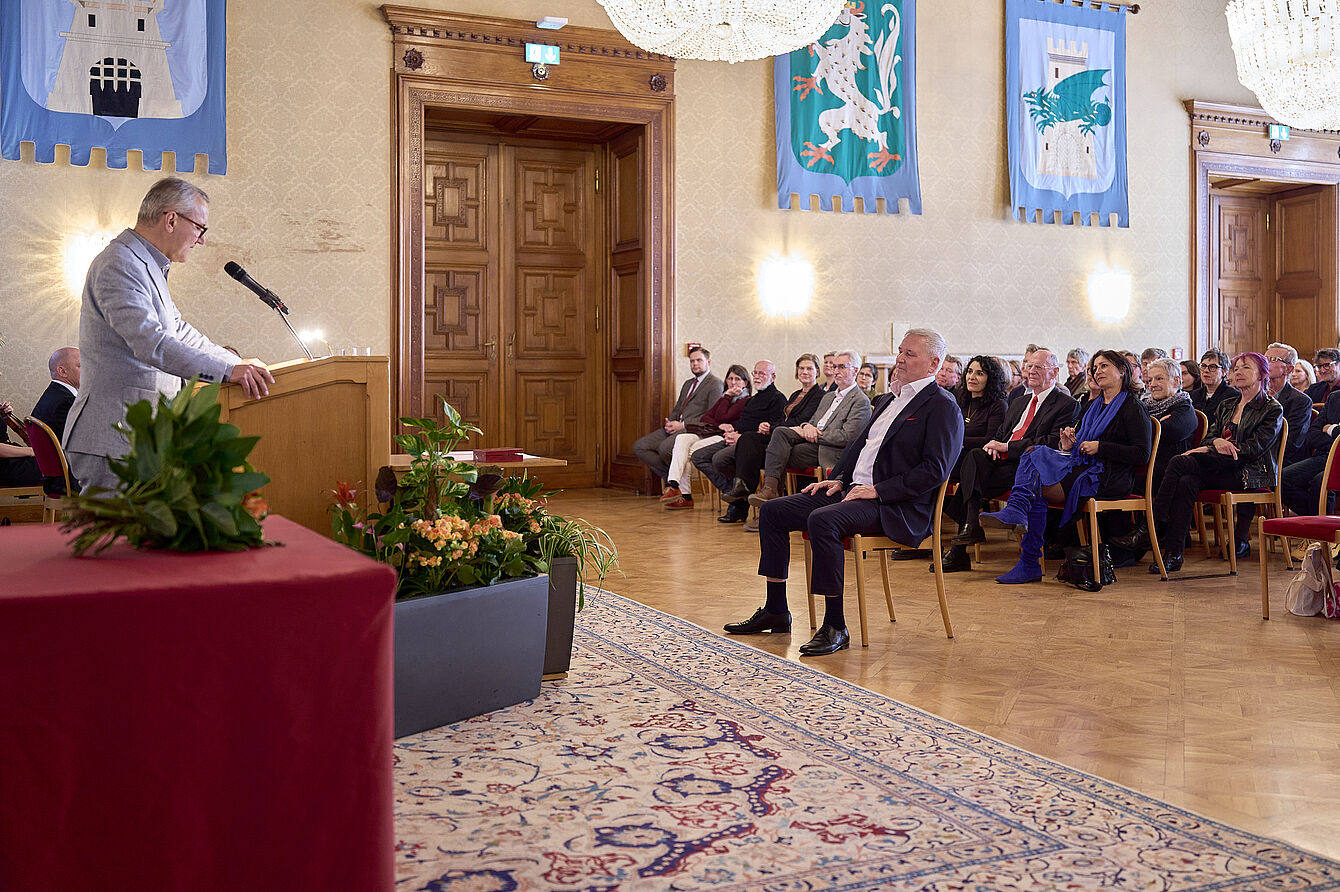 Redner an einem Rednerpult in einem festlichen Saal mit Publikum auf Stühlen und dekorativen Wappen an den Wänden