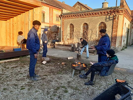 Mehrere Personen stehen und sitzen um ein kleines Feuer in einer Feuerschale vor einem Backsteingebäude