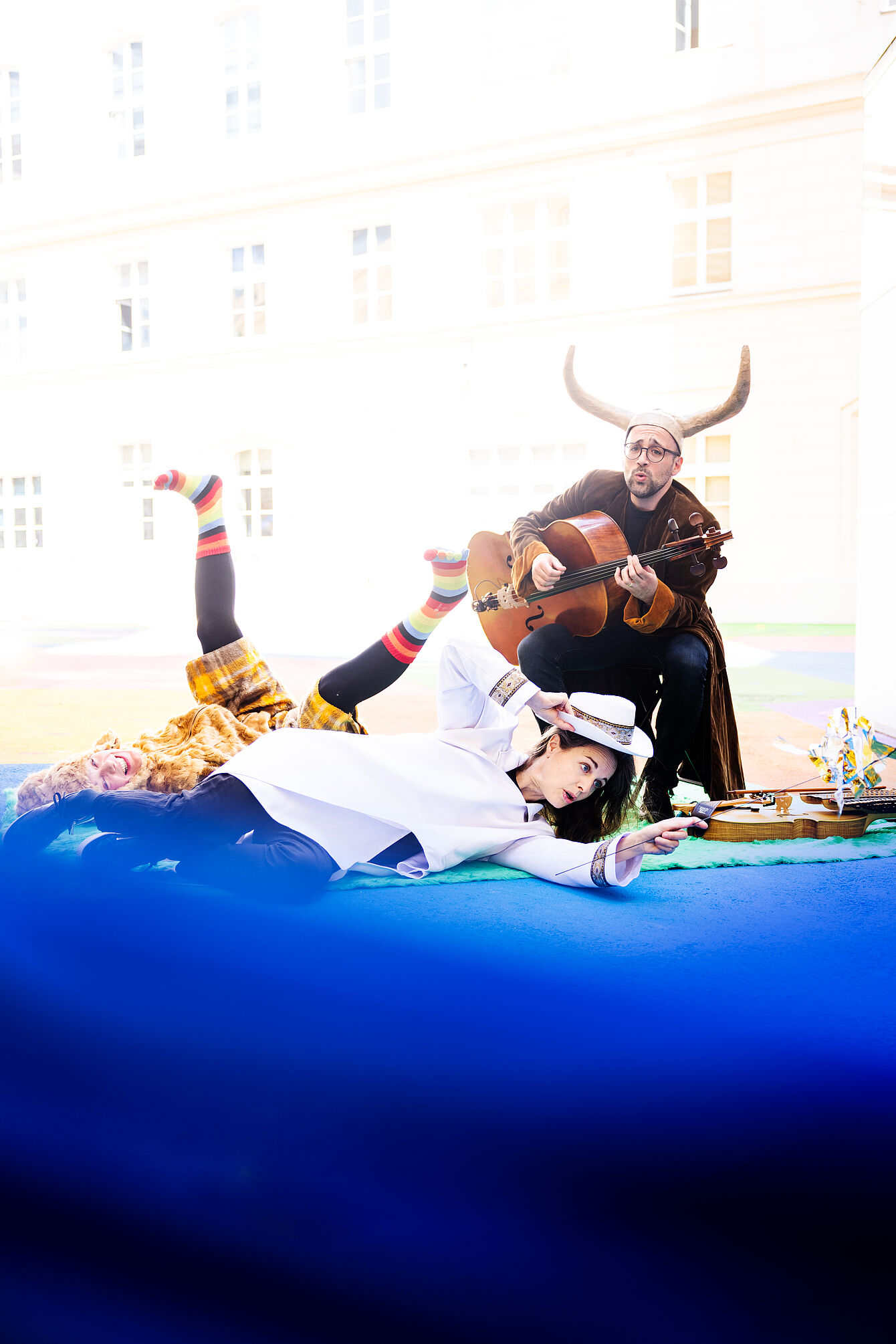 Person mit Hörnern und Umhang spielt Gitarre, daneben eine Person in bunter Kleidung mit hochgestreckten Beinen auf blauem Untergrund liegend.