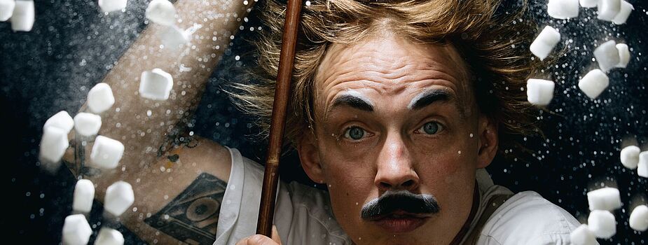 Person mit hochgekrempelten Ärmeln hält einen langen Stab, umgeben von fliegenden kleinen weißen Würfeln und weißem Pulver vor dunklem Hintergrund.