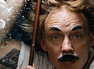 Person mit hochgekrempelten Ärmeln hält einen langen Stab, umgeben von fliegenden kleinen weißen Würfeln und weißem Pulver vor dunklem Hintergrund.