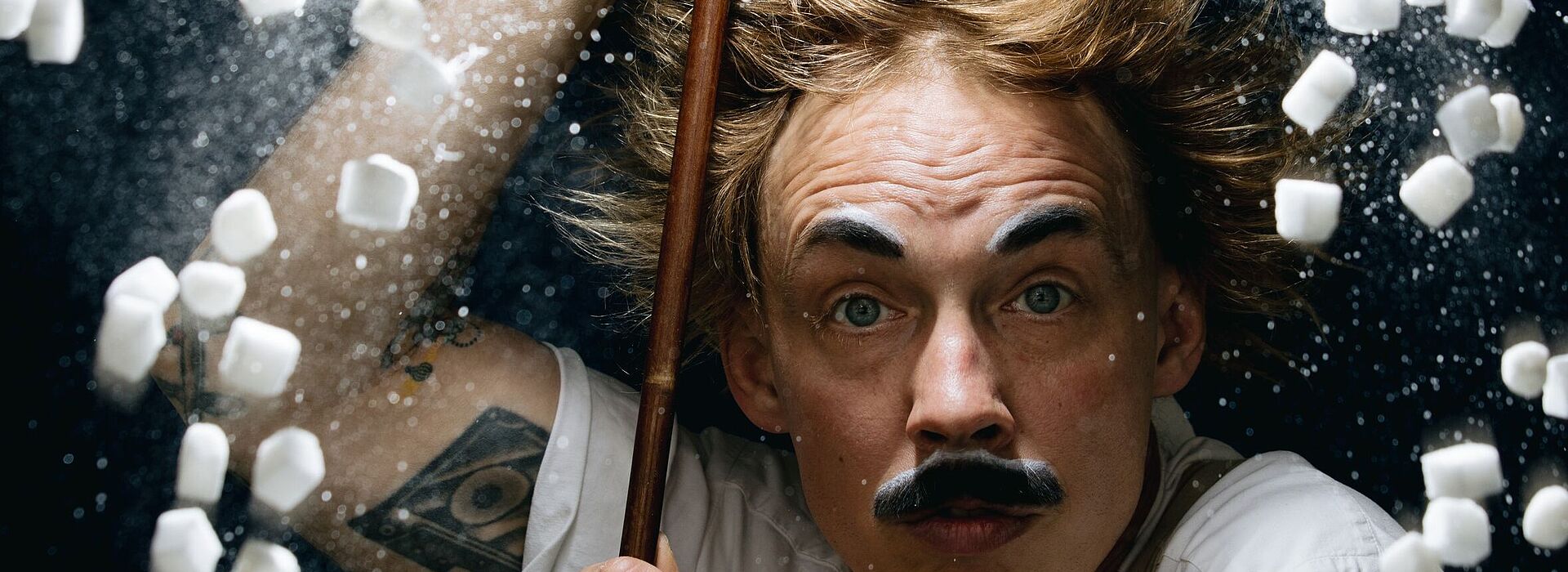 Person mit hochgekrempelten Ärmeln hält einen langen Stab, umgeben von fliegenden kleinen weißen Würfeln und weißem Pulver vor dunklem Hintergrund.