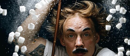 Person mit hochgekrempelten Ärmeln hält einen langen Stab, umgeben von fliegenden kleinen weißen Würfeln und weißem Pulver vor dunklem Hintergrund.