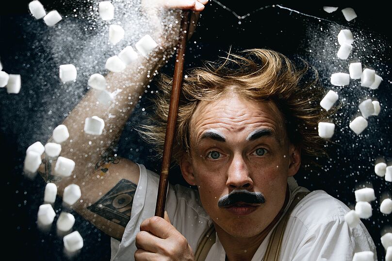 Person mit hochgekrempelten Ärmeln hält einen langen Stab, umgeben von fliegenden kleinen weißen Würfeln und weißem Pulver vor dunklem Hintergrund.