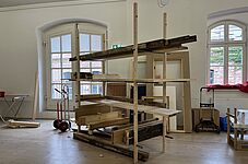 Eine Konstruktion aus Holzregalen steht in einem Raum mit großen Fenstern. Die Regale sind aus verschiedenen Holzarten und -farben, einige Bretter sind horizontal, andere vertikal angeordnet.