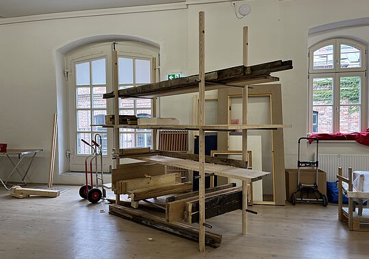Eine Konstruktion aus Holzregalen steht in einem Raum mit großen Fenstern. Die Regale sind aus verschiedenen Holzarten und -farben, einige Bretter sind horizontal, andere vertikal angeordnet.
