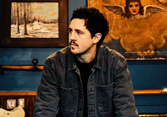 Ein Mann mit kurzen dunkelbraunen Haaren steht an einem Billard-Tisch und blickt auf die Seite, über ihm ein großer Luster mit Glasmalerei und hinter ihm eine blaue Wand mit unterschiedlichen Bildern