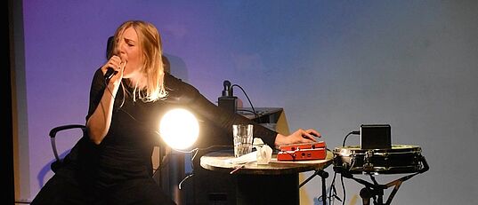 Person in schwarzer Kleidung sitzt auf Stuhl, hält Mikrofon und bedient mit der anderen Hand ein elektronisches Gerät auf einem Tisch mit Glas und Lampe