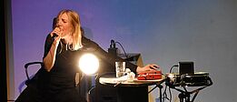 Person in schwarzer Kleidung sitzt auf Stuhl, hält Mikrofon und bedient mit der anderen Hand ein elektronisches Gerät auf einem Tisch mit Glas und Lampe